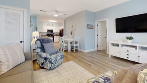 Een woonkamer met een bank, een gestreept fauteuil, een eettafel met stoelen, een televisie en een keuken op de achtergrond.