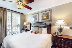 A hotel room with a large bed, a ceiling fan, a balcony with a view of buildings, and two framed pictures on the wall. - Inviting, Spacious Condo On Okaloosa Island Playground, 2 Hot Tubs (Fort Walton Beach)
