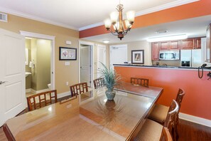 A dining area with a glass table, wooden chairs, a chandelier, and a kitchen in the background. - Inviting, Spacious Condo On Okaloosa Island Playground, 2 Hot Tubs (Fort Walton Beach)