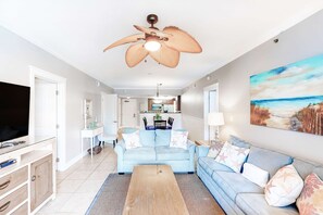 A spacious living room with a ceiling fan, a large flat-screen TV, a light blue sofa set, a wooden coffee table, and a beach-themed painting on the wall. - 2nd Floor Condo At Waterscape Steps To Beach Lazy River (Fort Walton Beach)