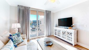 Uma sala de estar moderna com um sofá, uma mesa de centro de vidro, uma televisão em um móvel branco e uma varanda com cortinas.