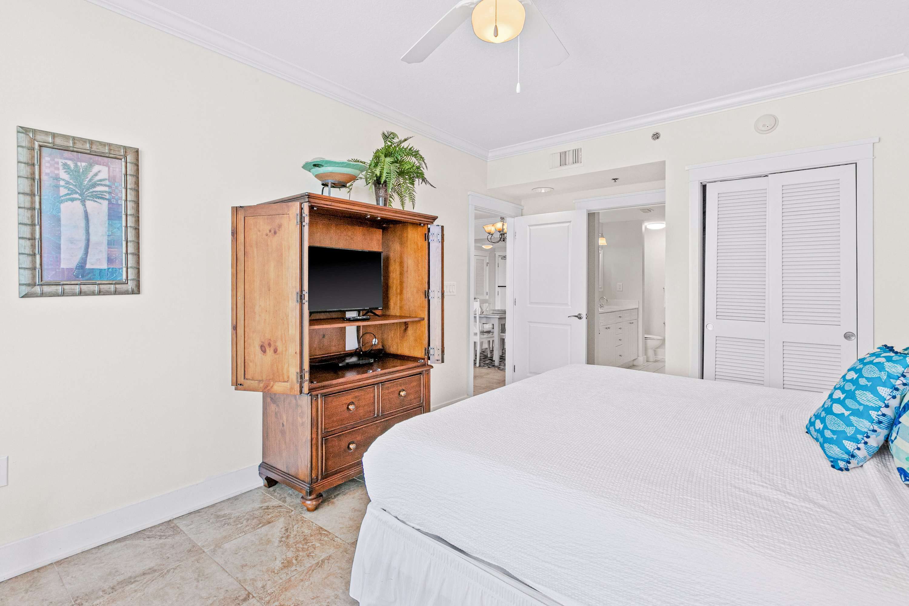 A bedroom with a large bed, a wooden dresser, a ceiling fan, and a door leading to a bathroom.