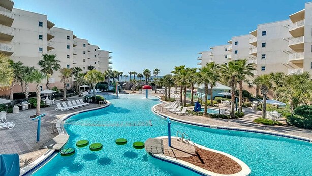 A swimming pool with lounge chairs and palm trees, surrounded by multi-story buildings. - 3rd Floor Spacious Condo At Waterscape Children's Playground, 2 Hot Tubs (Fort Walton Beach)