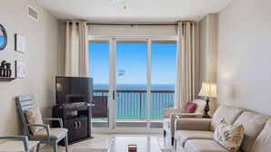Een kamer met een televisie, een bank, een stoel, een salontafel en een balkon met uitzicht op de oceaan.