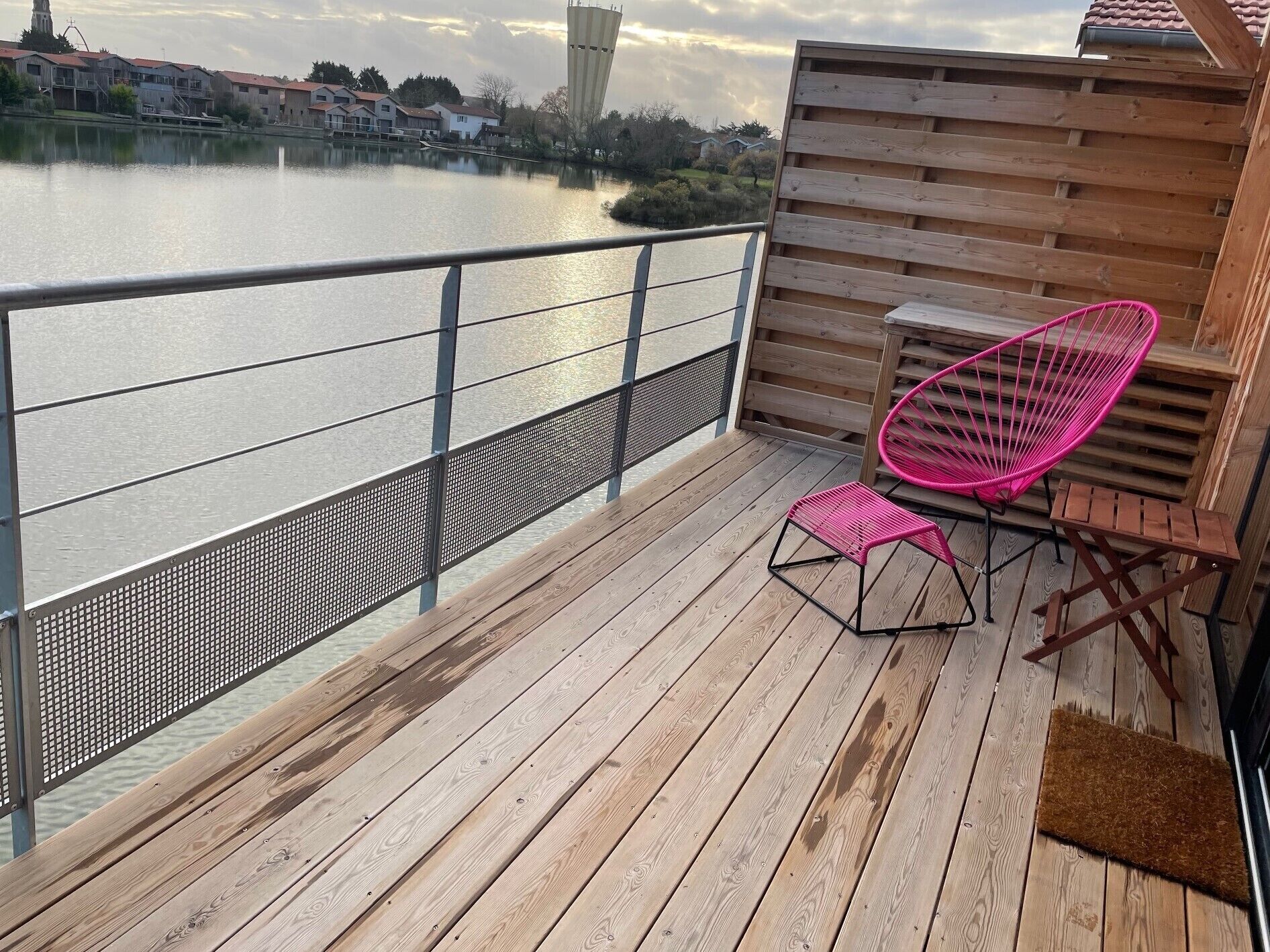 A wooden deck with a metal railing overlooking a body of water, a pink chair, and a small table.