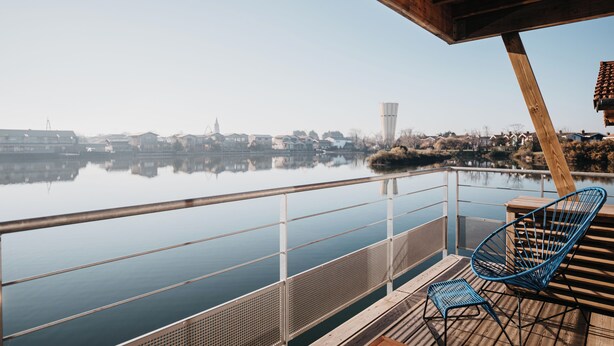 A wooden deck with a railing overlooking a calm body of water, a chair, and a small table. - LES 12 DE LARROS (Gujan-Mestras)