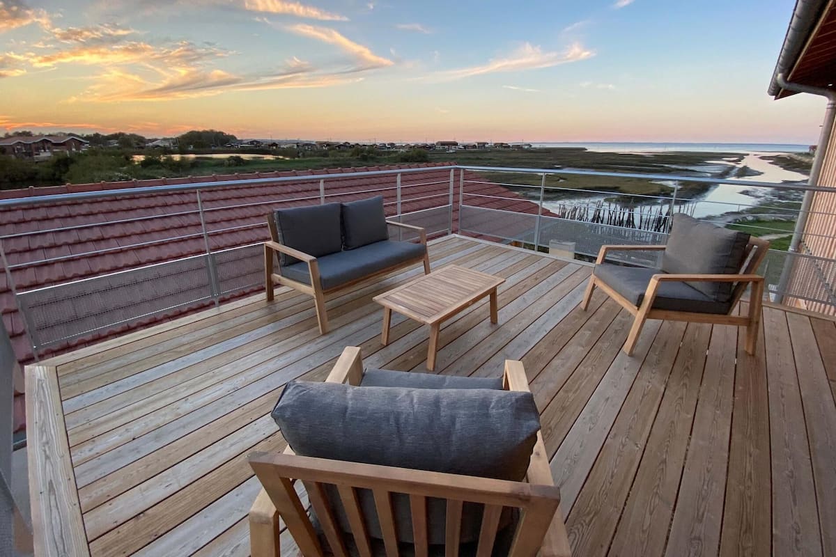 A wooden deck with outdoor furniture, a railing, and a view of a water body and distant landscape.