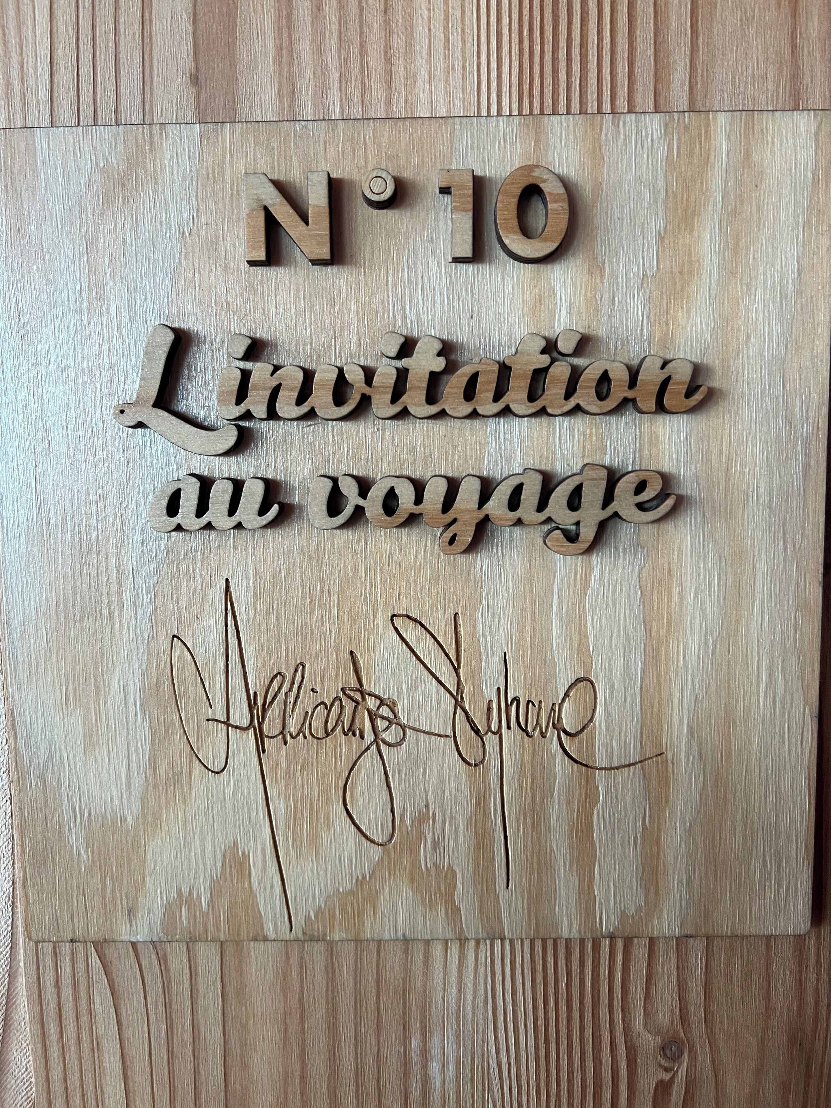 A wooden sign with the text "N°10 Invitation au voyage" and a signature.