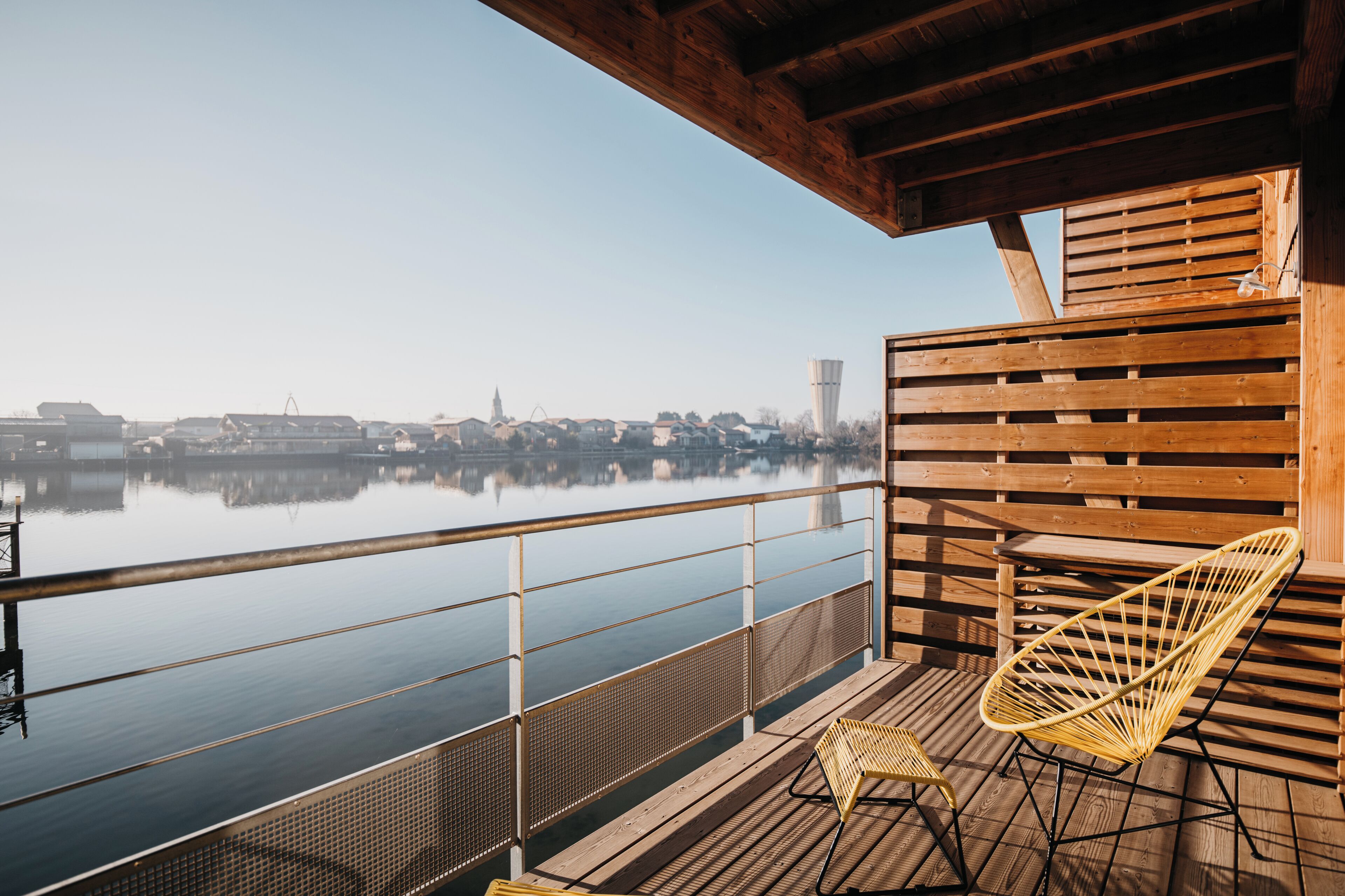 Una terraza de madera con barandilla que da a una masa de agua tranquila, una silla y una pequeña mesa.