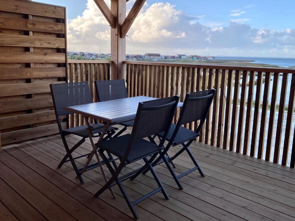 Um deck de madeira com mesa e cadeiras, com vista para uma praia e edifícios ao longe.
