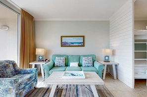 Un salon avec un canapé de couleur turquoise, deux coussins à motifs, une table basse avec un plateau et un tableau représentant une scène de plage.