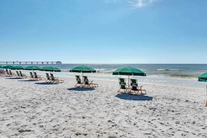 Ein Strand mit Reihen von Liegestühlen und grünen Sonnenschirmen, ein Steg im Hintergrund und ein klarer Himmel.