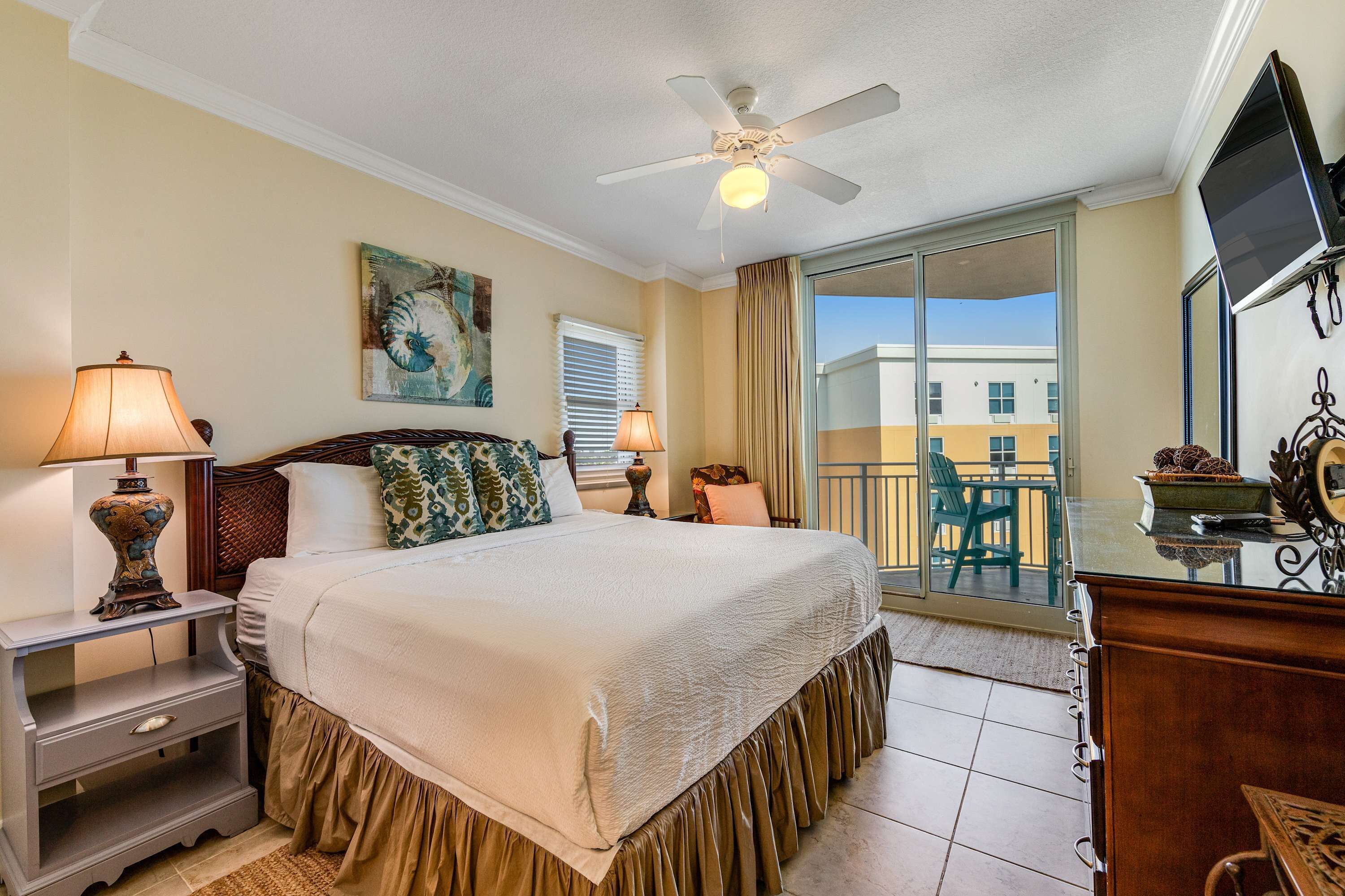 A hotel room with a bed, nightstands, a ceiling fan, a TV, and a balcony with a view of buildings.
