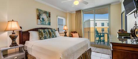 A hotel room with a bed, nightstands, a ceiling fan, a TV, and a balcony with a view of buildings.