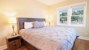 Un dormitorio con una cama grande, dos mesitas de noche con lámparas, una ventana con vistas a árboles y suelo de madera.