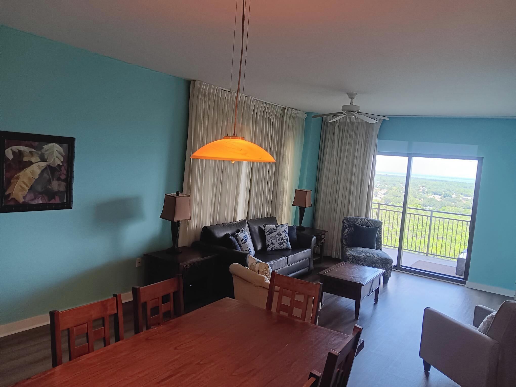 Sala de estar com sofá, mesa de jantar e cadeiras. Há uma janela grande com vista para área verde.