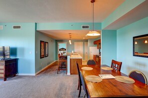 A dining area with a wooden table, chairs, and a kitchen in the background. - Beautiful corner condo with Gulf & bay views, balcony, pool & hot tub (Panama City Beach)