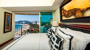 A hotel room with a bed, a painting, a view of the beach and cityscape, and a balcony with a railing. - Bay View Grand Residencial 11th floor (Ixtapa Zihuatanejo)