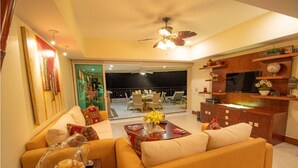 A well-lit living room with a sofa, coffee table, and a TV on a wooden cabinet. There is a balcony with chairs and a table. - Bay View Grand Residencial 11th floor (Ixtapa Zihuatanejo)
