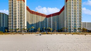 Ein Hochhaus-Hotel mit mehreren Balkonen, ein Strand im Vordergrund und Palmen.