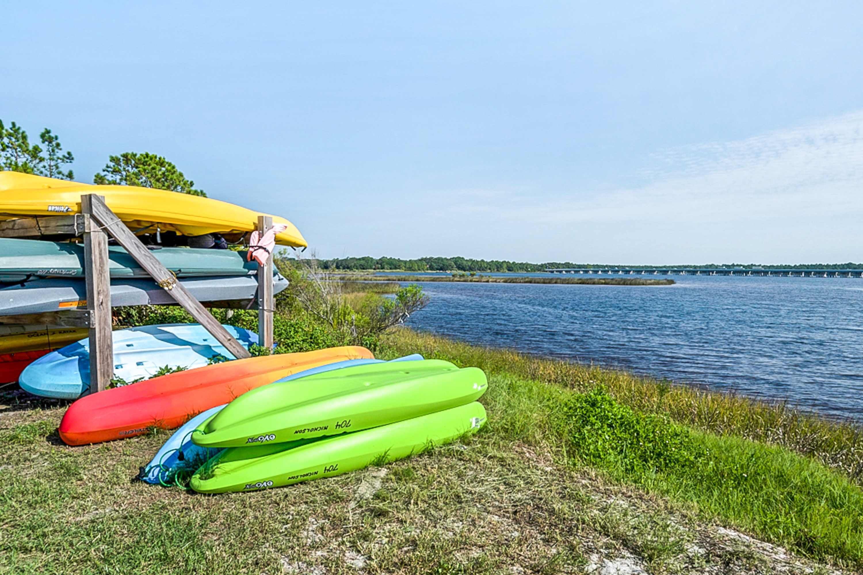 A row of kayaks and a canoe on a grassy shore by a calm lake with a bridge in the distance.