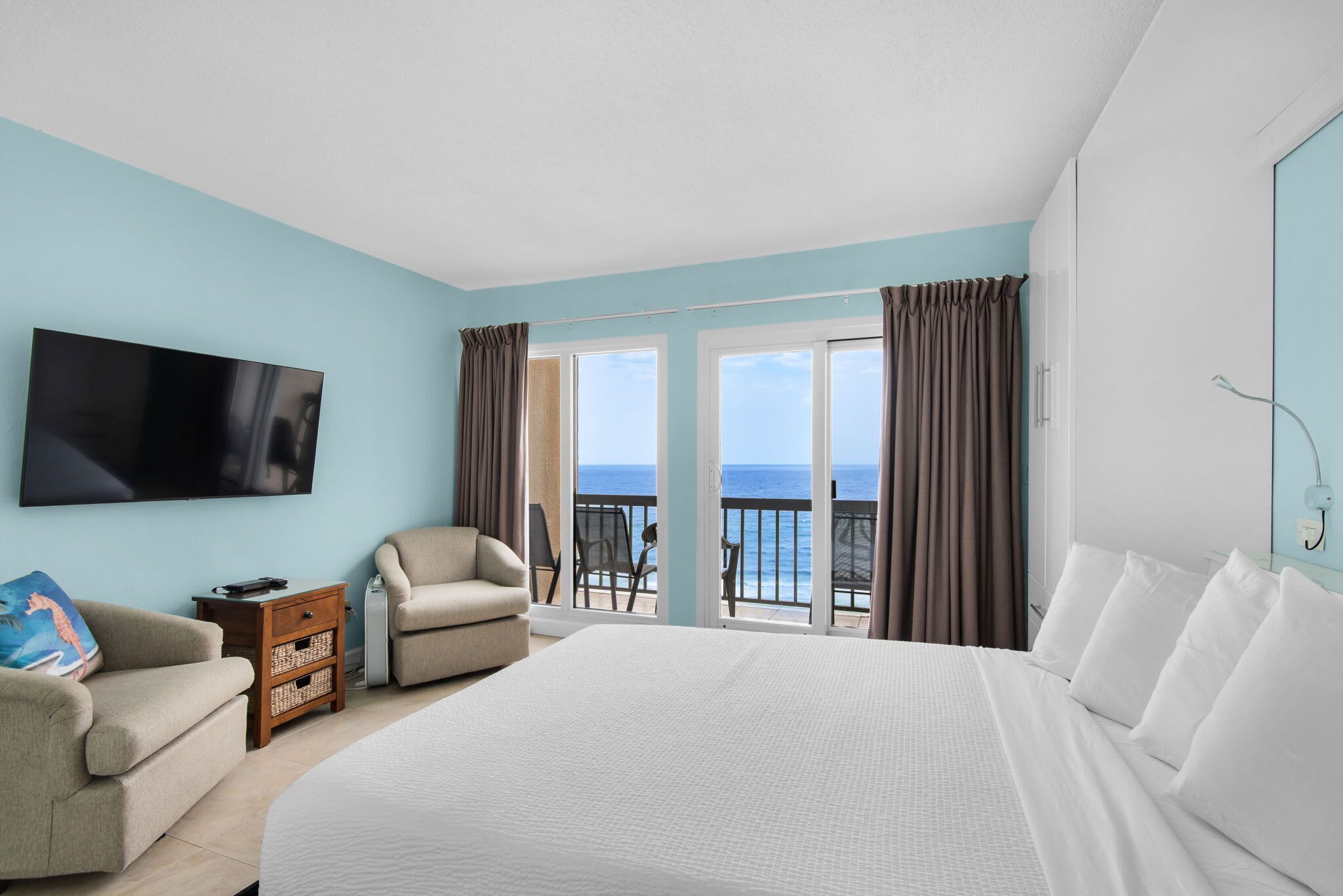 Une chambre d’hôtel avec un grand lit, un téléviseur à écran plat, une chaise, une petite table et une vue sur l’océan depuis les portes du balcon.