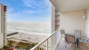 Een balkon met uitzicht op strand en zee, een tafel met stoelen, en een gebouw met meerdere verdiepingen en balkons.