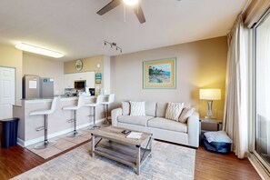 Ein modernes Wohnzimmer mit einer Couch, einem Couchtisch und Barhocker. Es gibt eine Küchenzeile mit Kühlschrank und Mikrowelle. Ein Gemälde hängt an der Wand, und eine Lampe steht auf einem Beistelltisch.