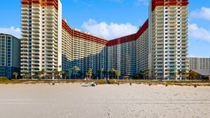 Un hotel frente a la playa con varios edificios altos, una playa de arena en primer plano y palmeras.