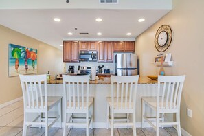A kitchen with wooden cabinets, a stainless steel refrigerator, and a painting of palm trees on the wall. - Kid-friendly beachfront condo with seasonal beach service, pools & hot tub (Panama City Beach)