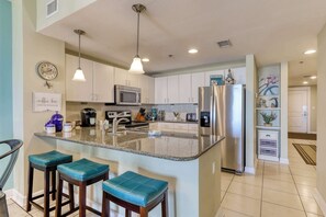 A modern kitchen with a central island, stainless steel appliances, and teal bar stools. - Oceanfront getaway with views, large balcony, W/D, pool & hot tub (Panama City Beach)