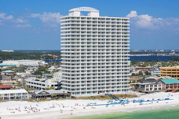 A tall hotel with multiple floors, a beach in the foreground, and a view of the ocean and other buildings in the background. - Beachfront, seafoam sanctuary with furnished balcony, pools, & gym (Panama City Beach)