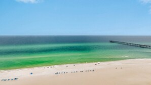 Una playa con un muelle que se adentra en el agua, cielos despejados y mar en calma.