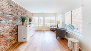 A modern living room with a brick wall, wooden flooring, a grey sofa, a small round table, and a cabinet with drawers. - Updated townhome with grassy yard, grill, open layout, W&D, & AC (Perdido Key)