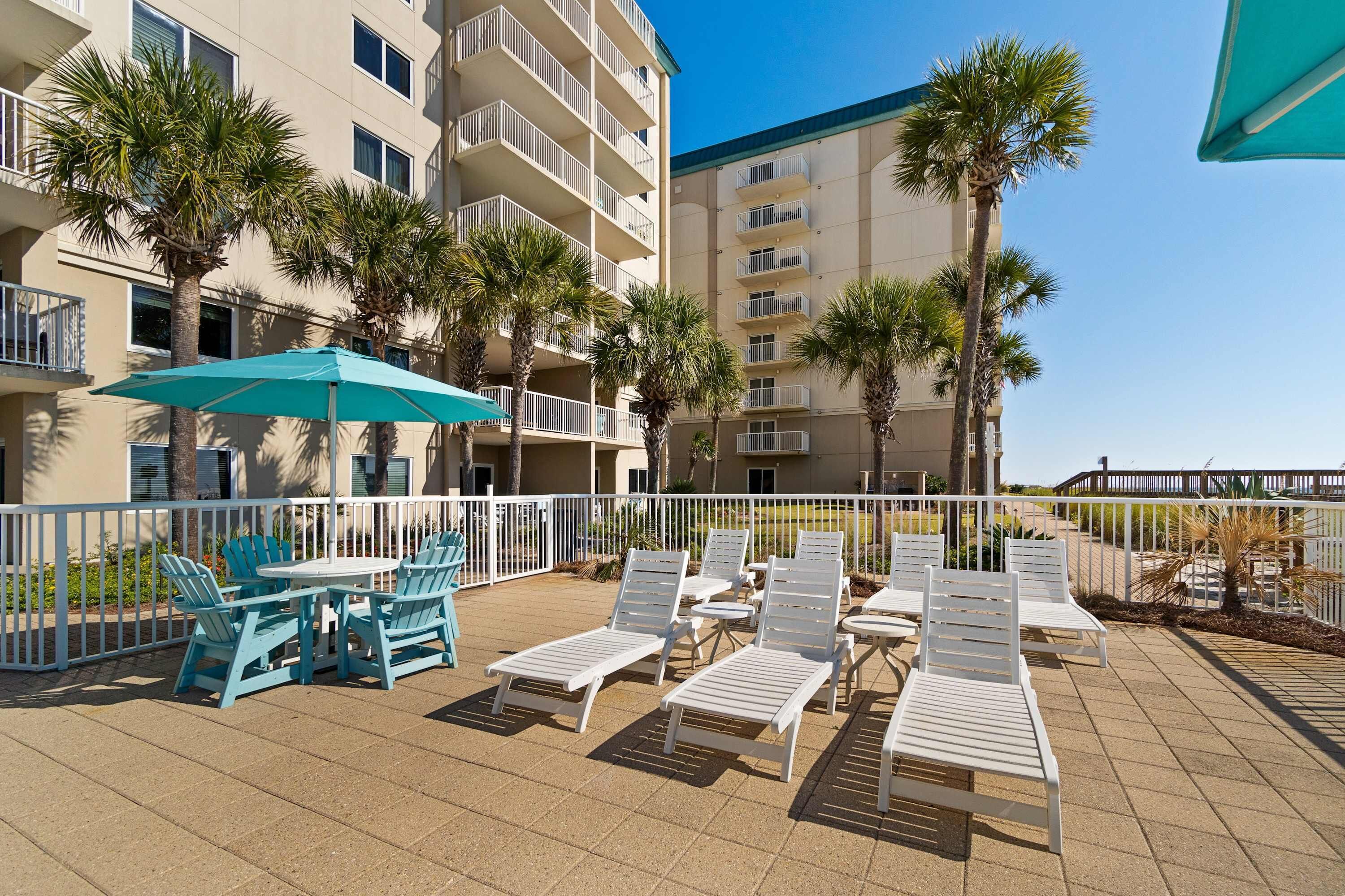 A rooftop patio with white lounge chairs and a table, surrounded by a white fence, with palm trees and a building in the background.