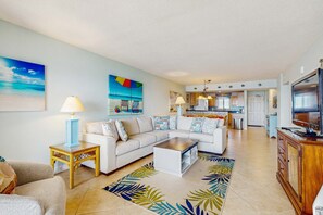 A living room with a sofa, coffee table, and a TV. There is a kitchen in the background. - Gorgeous Oceanfront condo with washer/dryer, Gulf-front balcony & pool/hot tub (Perdido Key)
