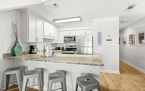 Eine moderne Küche mit weißen Schränken, einer Marmorarbeitsplatte und Edelstahlgeräten. Es gibt drei Barhocker und eine Vase mit Ästen.