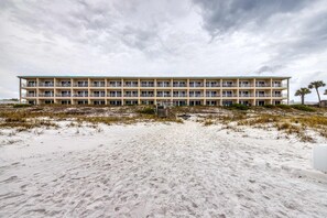 Ein mehrstöckiges Hotel mit flachem Dach und Balkonen, an einem Sandstrand mit spärlicher Vegetation gelegen.