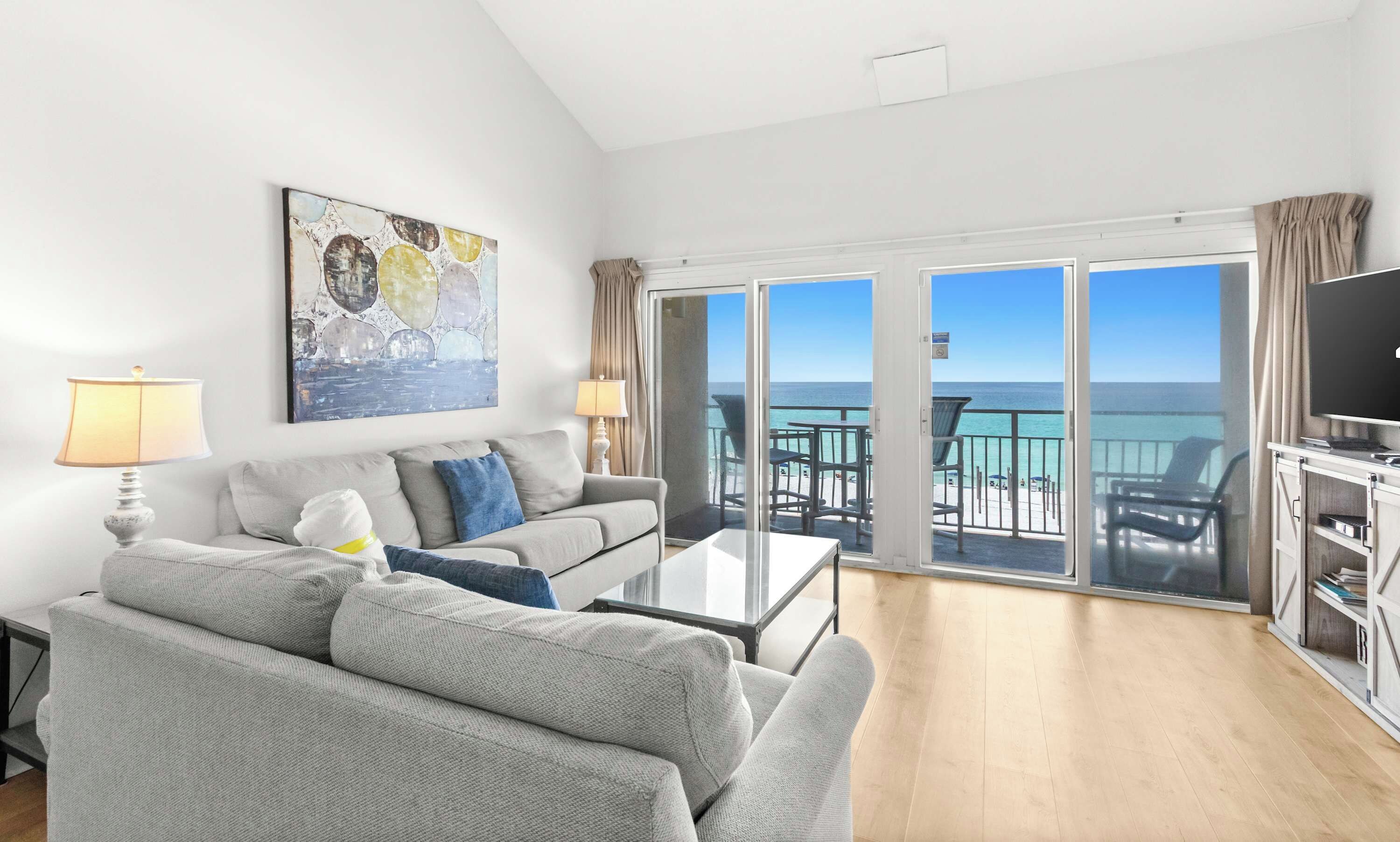 A modern living room with a sofa, a coffee table, and a painting on the wall. There is a balcony with a view of the ocean.