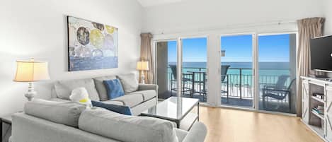 A modern living room with a sofa, a coffee table, and a painting on the wall. There is a balcony with a view of the ocean.