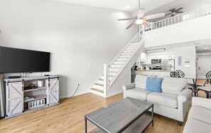 A modern living room with a flat-screen TV, a sofa, a coffee table, and a staircase leading to a kitchen area. - Stunning third-floor condo with balcony, washer&dryer & pool (Destin)