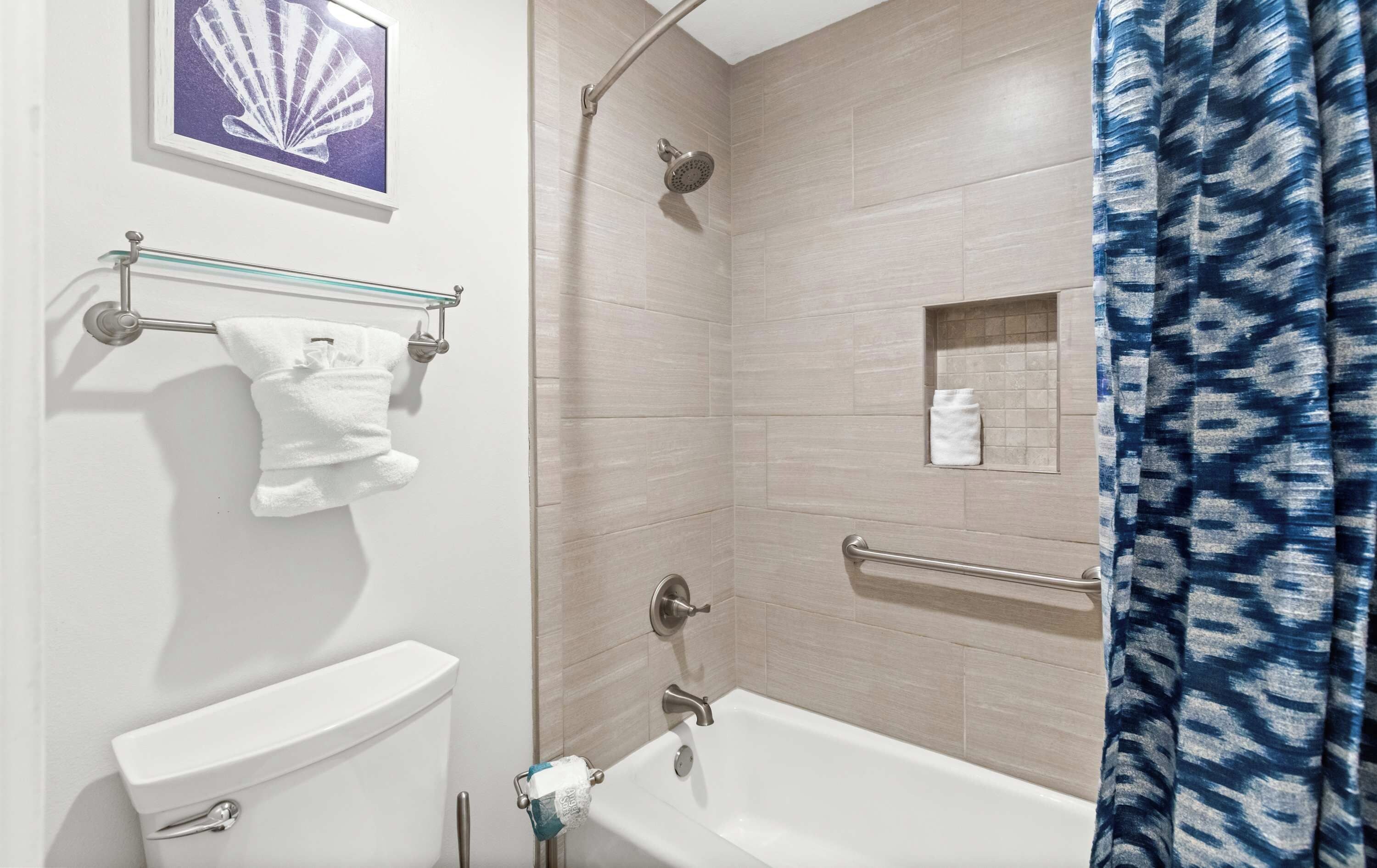 A bathroom with a toilet, a shower with a handheld showerhead, a towel rack with a white towel, and a framed picture on the wall.