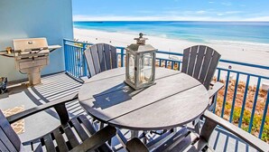 A balcony with a round table and chairs, a grill, and a lantern. The balcony overlooks a beach with clear blue water and a sandy shore. - Gulf-front condo with balcony, beach access, hot tub, pool & gym (Panama City Beach)