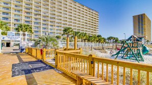 Ein hölzerner Steg führt zu einem Strandbereich mit Spielplatz, Palmen und einem großen Hotel im Hintergrund.