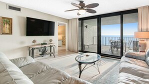 Ein modernes Wohnzimmer mit Flachbildfernseher, Deckenventilator, Glastisch und einem Balkon mit Meerblick.