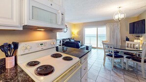 A kitchen with a white stove, microwave, and cabinets, a dining area with a table and chairs, and a view of the outside through a large window. - Gulf-front condo with balcony, W/D, direct beach access, pools, hot tub & gym (Panama City Beach)