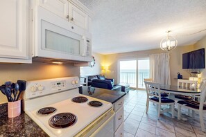 A kitchen with a white stove, microwave, and cabinets, a dining area with a table and chairs, and a view of the outside through a large window. - Gulf-front condo with balcony, W/D, direct beach access, pools, hot tub & gym (Panama City Beach)