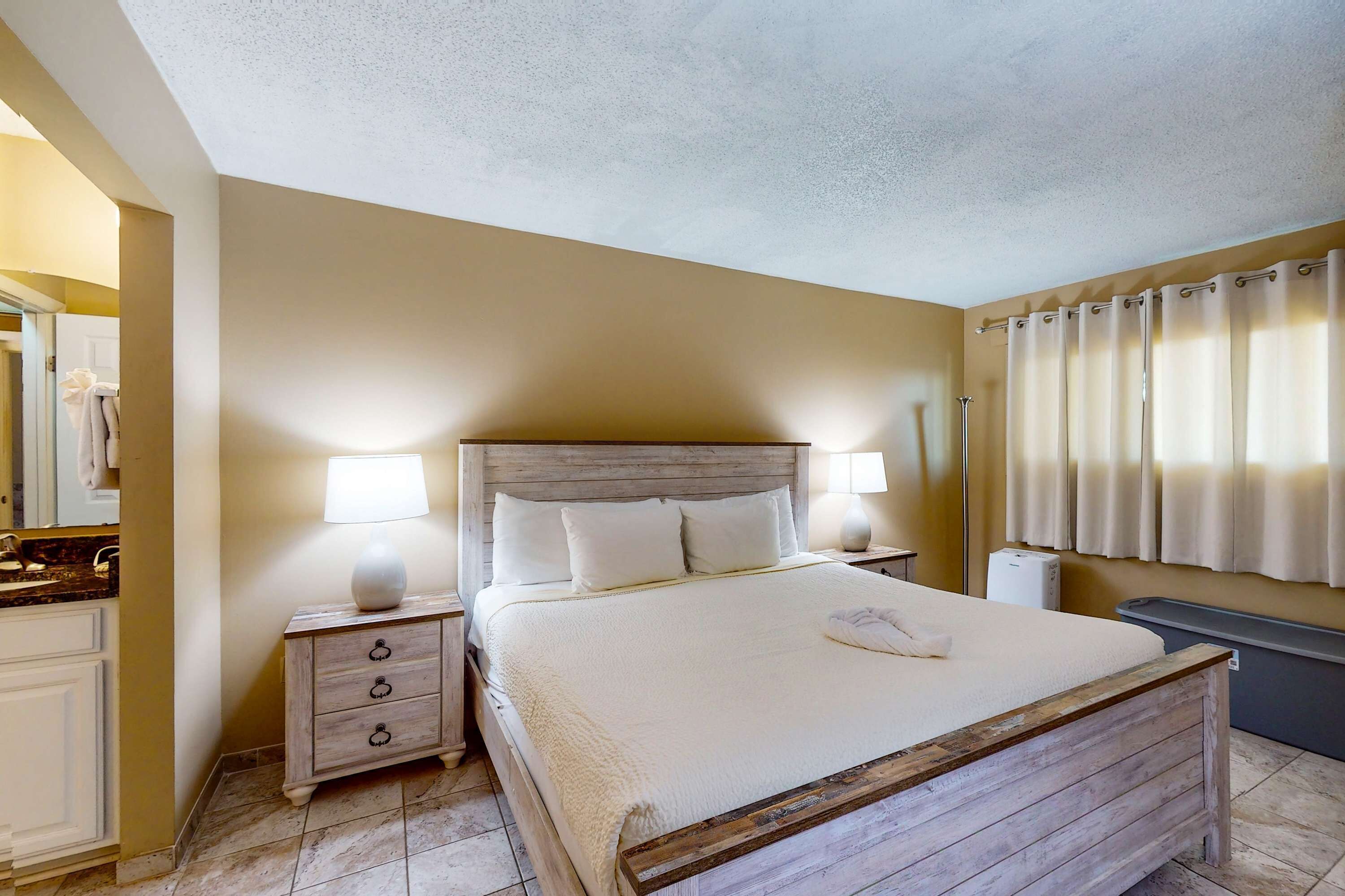 Quarto com cama de madeira, roupa de cama branca, mesa de cabeceira com abajur, cômoda e banheiro com cortina de chuveiro.
