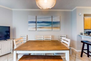 A dining area with a wooden table, white chairs, a bench, and a large abstract painting on the wall. - Lovely condo with pool, hot tub, ocean views & pickleball (Santa Rosa Beach)