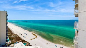 A beach view from a high-rise building with a clear sky, a pier extending into the ocean, and a sandy beach with umbrellas and lounge chairs. - High-rise condo at beachfront resort w/ pools, hot tubs & arcade (Panama City Beach)